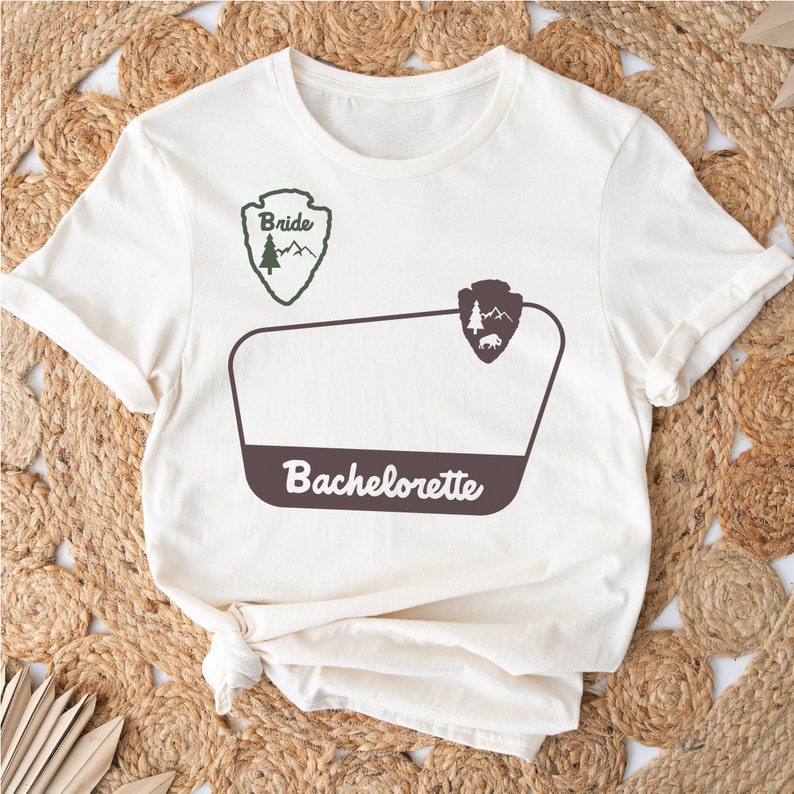 May include: White t-shirt with a brown and green graphic of a national park sign. The sign says "Bride" and "Bachelorette" with a small tree and mountain design.