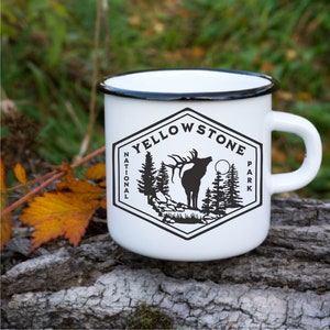 May include: White enamel mug with a black rim and handle. The mug features a black and white graphic of an elk in a forest with the text "Yellowstone National Park".