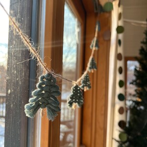 May include: A string of teal blue macrame Christmas tree ornaments hanging in a window. The ornaments are strung on twine and are hanging in front of a wooden window frame.