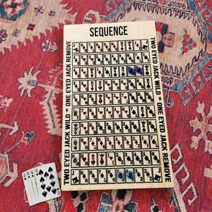 May include: A wooden game board with the word "SEQUENCE" printed on it. The board has a grid of squares with numbers and symbols. There are two blue game pieces on the board. The text "TWO EYED JACK WILD * ONE EYED JACK REMOVE" is printed on the side of the board.