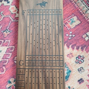 May include: A wooden horse racing game board with a black engraved design. The board has a track with 12 numbered spaces and a betting area with dollar amounts: $1, $0.75, $0.50, and $0.25.
