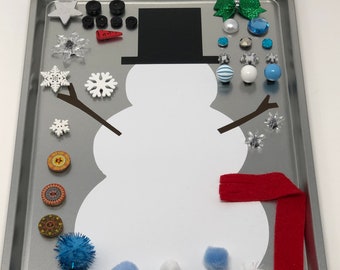 May include: A white snowman craft activity with a black top hat, red scarf, and various embellishments including snowflakes, buttons, pom-poms, and beads.