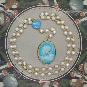 May include: A white pearl necklace with a gold chain and a blue and white oval pendant featuring a mother and child. The pendant is attached to a blue heart charm.