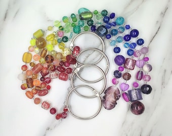 May include: A collection of colorful beaded jewelry arranged in a rainbow gradient. The beads are various shapes and sizes, with a mix of clear and opaque finishes. Silver metal rings are in the center of the arrangement.