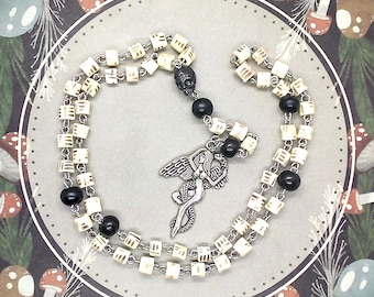 May include: A necklace with off-white cube beads with etched designs, and black beads. A silver pendant of a mermaid with wings and a skull bead are central to the design. The necklace is set against a whimsical mushroom background.