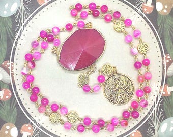 May include: A necklace featuring a gold-tone chain with pink beads and circular tree-of-life charms. The piece includes a large, faceted, oval-shaped magenta pendant and a round gold-tone medallion depicting a figure with raised arms.