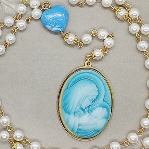 May include: A white pearl rosary necklace with a gold chain and a blue heart charm. The pendant features a gold oval frame with a blue and white image of the Virgin Mary and baby Jesus.