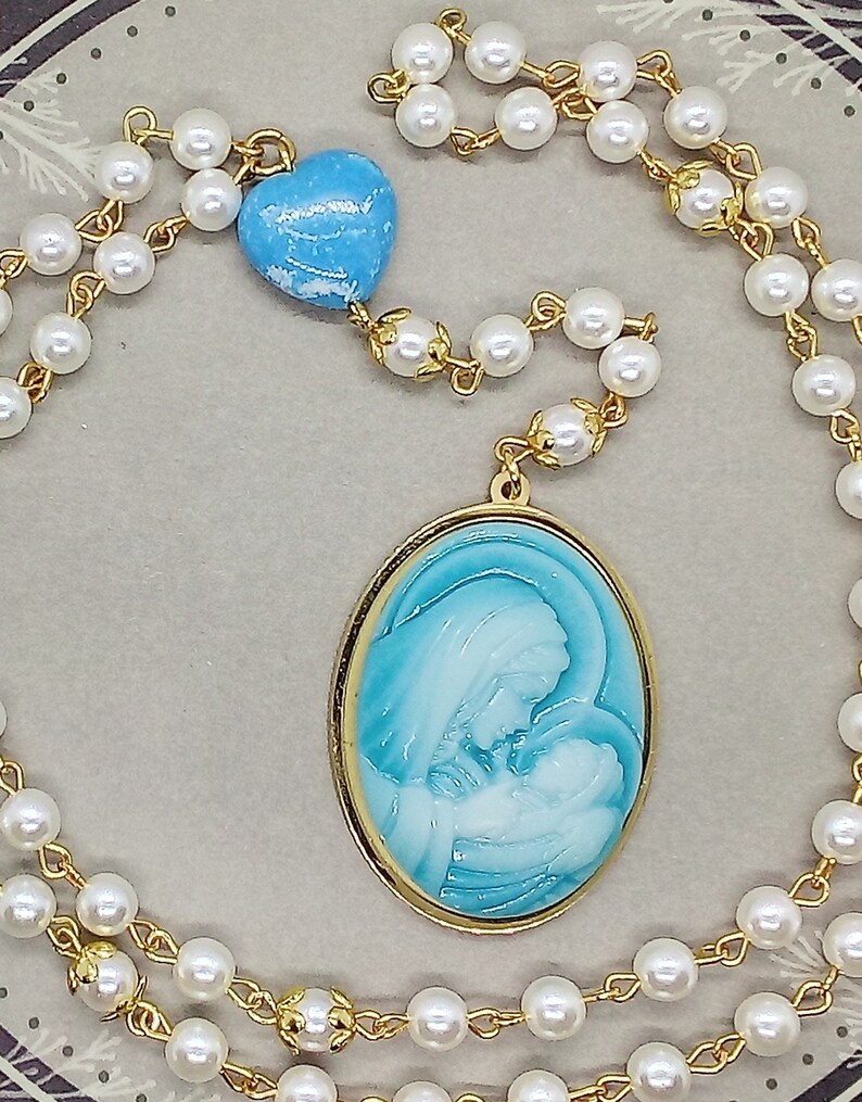 May include: A white pearl rosary necklace with a gold chain and a blue heart charm. The pendant features a gold oval frame with a blue and white image of the Virgin Mary and baby Jesus.