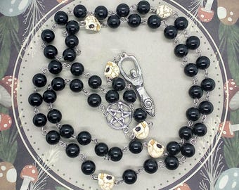 May include: A black beaded necklace with skull accents and silver-toned pendants. The necklace features a pentagram and a stylized female figure pendant. The beads are round and glossy, and the skulls are off-white.