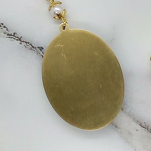 May include: A gold oval pendant with a smooth, shiny surface. The pendant is attached to a chain with white pearl beads.