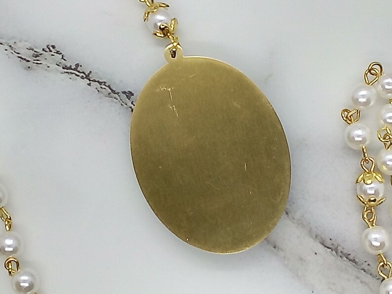 May include: A gold oval pendant with a smooth, shiny surface. The pendant is attached to a chain with white pearl beads.