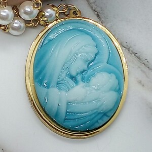 May include: A gold oval pendant with a blue and white cameo of the Virgin Mary holding a baby Jesus. The pendant is attached to a white pearl necklace.