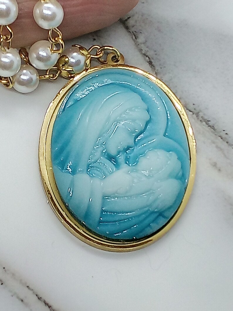 May include: A gold oval pendant with a blue and white cameo of the Virgin Mary holding a baby Jesus. The pendant is attached to a white pearl necklace.