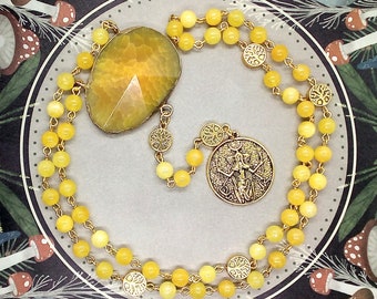 May include: A necklace featuring a large, faceted yellow stone pendant and a round gold-toned medallion with an embossed figure. The chain consists of small, round yellow beads interspersed with circular gold-toned charms depicting tree motifs.