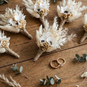 Op de afbeelding: Een verzameling handgemaakte corsages met witte anemoonbloemen, gedroogde grassen en met touw omwikkelde stelen. Twee gouden trouwringen, waarvan één met een diamant, liggen op een rustiek houten oppervlak. Eucalyptustakjes.