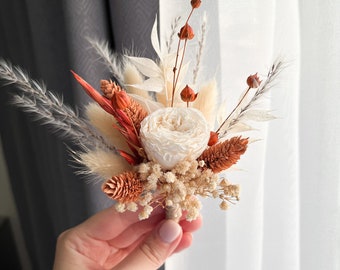 Puede incluir: Un prendido hecho a mano con una rosa blanca como punto focal, rodeado de flores secas en tonos naranja, beige y crema. El arreglo incluye pequeñas piñas y acentos plumosos, todo atado con hilo.