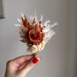 Puede incluir: Un pequeño ramo de flores secas en tonos marrones y blancos. El ramo presenta una sola rosa naranja, hierba de la pampa blanca y otras hojas secas.