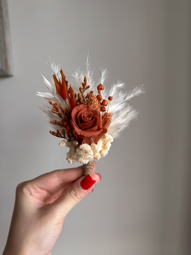 Puede incluir: Un peque&ntilde;o ramo de flores secas en tonos marrones y blancos. El ramo presenta una sola rosa naranja, hierba de la pampa blanca y otras hojas secas.