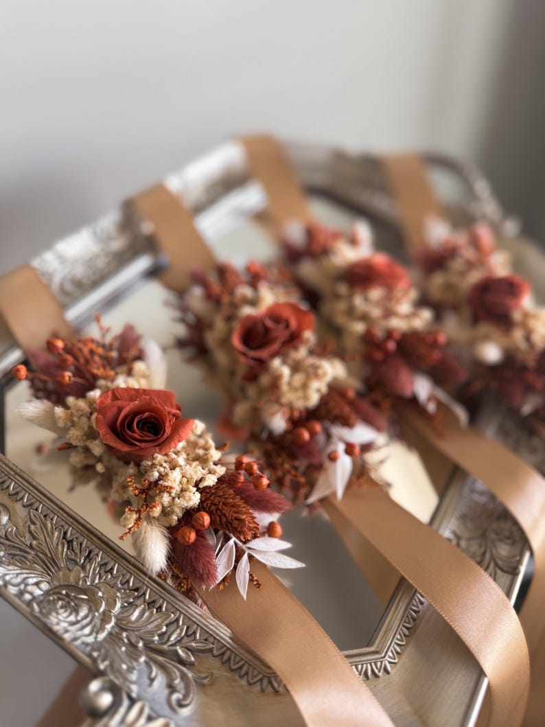 Puede incluir: Cuatro corsages de mu&ntilde;eca de cinta de sat&eacute;n marr&oacute;n con flores secas en tonos naranja, blanco y marr&oacute;n. Los corsages est&aacute;n dispuestos en una bandeja de plata.