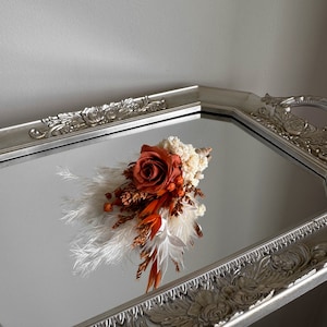 Puede incluir: Un peque&ntilde;o ramo de flores secas en tonos naranja y blanco, incluyendo una sola rosa naranja, exhibido en una bandeja plateada con un dise&ntilde;o ornamentado.