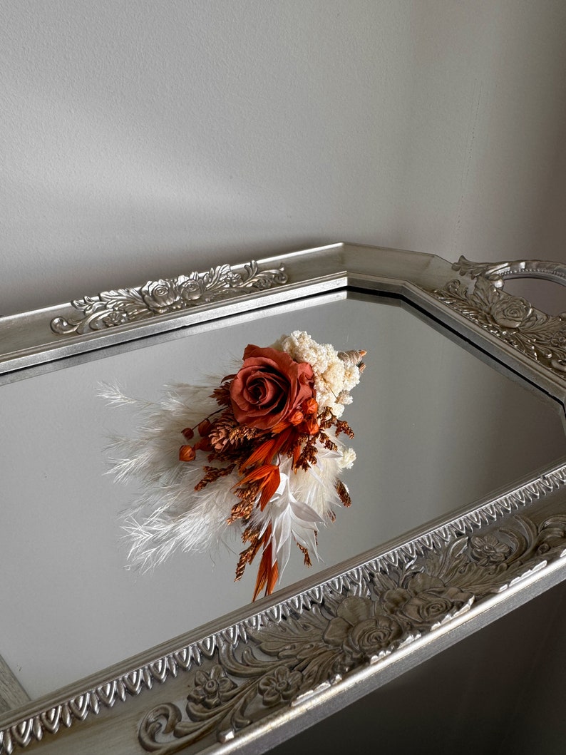 Puede incluir: Un peque&ntilde;o ramo de flores secas en tonos naranja y blanco, incluyendo una sola rosa naranja, exhibido en una bandeja plateada con un dise&ntilde;o ornamentado.