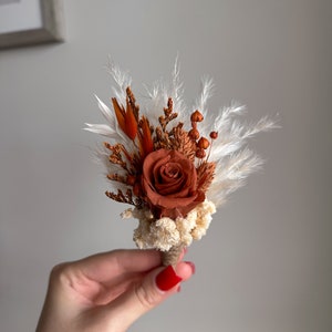 Puede incluir: Un peque&ntilde;o ramo de flores secas en tonos marrones y blancos, con una sola rosa naranja.