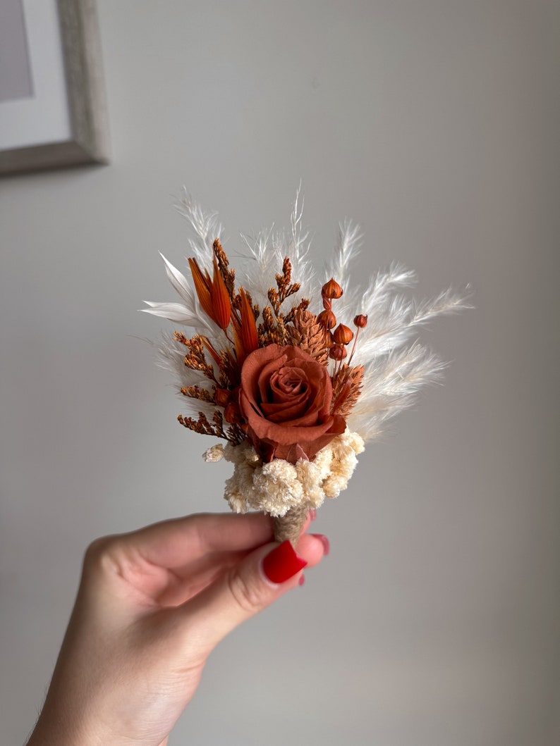Puede incluir: Un peque&ntilde;o ramo de flores secas en tonos marrones y blancos, con una sola rosa naranja.