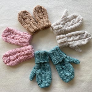 Puede incluir: Tres pares de manoplas tejidas a mano dispuestas en círculo. Los pares son de color rosa suave, beige claro y azul apagado. Cada manopla presenta un patrón de trenzas vertical en el dorso y un puño acanalado en la abertura de la muñeca.