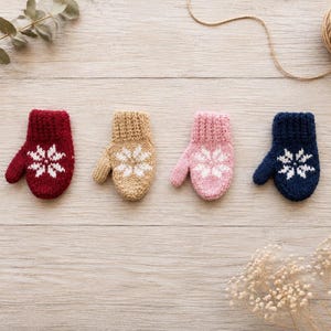 Könnte beinhalten: Vier kleine gestrickte Fäustlinge in einer Reihe, jeder mit einem gerippten Bündchen und einem weißen Schneeflockenmuster. Die Fäustlinge sind in Dunkelrot, Beige, Hellrosa und Marineblau gehalten und weisen eine einheitliche Strickstruktur auf.