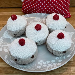 May include: Five knitted cupcakes sit on a grey plate. Each cupcake features a white top, a grey base, and a red raspberry. The plate has a floral pattern. A red and white polka dot cushion is in the background.