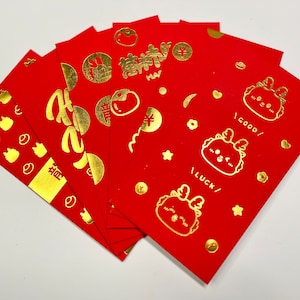 May include: A stack of red envelopes with gold foil designs. The envelopes feature various graphics, including cartoon characters, Chinese characters, and other festive symbols. The envelopes are likely for a holiday or special occasion.