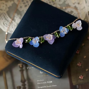 Puede incluir: Un delicado collar de oro con pequeños colgantes de flores de color morado, azul y verde. El collar se muestra en una caja de terciopelo azul.