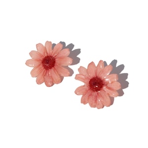 Puede incluir: Dos delicados pendientes en forma de margarita rosa claro con un centro rojo más oscuro. Los pendientes están sobre un fondo blanco, con sombras que sugieren una fuente de luz natural. El diseño floral añade un toque de elegancia inspirada en la naturaleza.