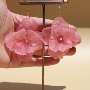 Puede incluir: Pendientes florales rosas con ganchos dorados. Cada pendiente presenta un grupo de pétalos delicados y translúcidos con una pequeña perla en el centro. Los pendientes se exhiben en un soporte de color cobre.