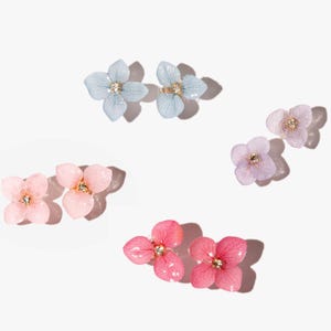 Könnte beinhalten: Ein Set mit sechs zarten, blumenförmigen Ohrringen in drei Farben: hellblau, rosa und Lavendel. Jeder Ohrring hat einen kleinen Strassstein in der Mitte.
