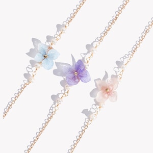 Könnte beinhalten: Drei zarte goldene Kettenarmbänder mit Perlenakzenten und einem einzelnen gepressten Blumenanhänger in Blau, Lila und Rosa.