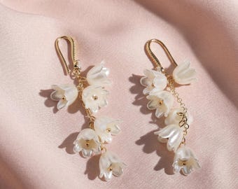 May include: A pair of gold-tone drop earrings featuring clusters of white, bell-shaped flowers. The earrings have a delicate chain and hook closure, with small gold accents in the center of each flower. The earrings are displayed on a soft, pink fabric.