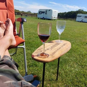 May include: A wooden table with a personalized inscription reading "Carl & Jen Home is where you park it."  Two glasses, one with red wine and the other with water, sit on the table.  The table is on a grassy lawn with a camper van in the background.