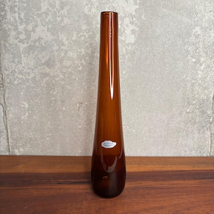 Pode incluir: Um vaso de vidro cor de âmbar, alto e esguio, com uma silhueta lisa e cónica. O vaso apresenta um pequeno autocolante branco de forma oval na superfície frontal com o texto "RUENKO HANDCRAFT" impresso a preto, indicando a sua origem e design.