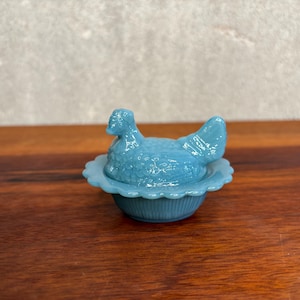 Könnte beinhalten: Eine hellblaue Glasschale mit einem Deckel in Form einer sitzenden Henne. Der Boden der Schale hat einen gewellten Rand und vertikale Rillen, während der Hennen-Deckel ein strukturiertes Federmuster auf Körper und Schwanz aufweist.