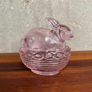 Könnte beinhalten: Eine durchscheinende, hellrosa Figur aus Pressglas, die einen Hasen in einem geflochtenen Korb darstellt. Der Hase weist eine texturierte Fellzeichnung und lange Ohren auf, während der Korb einen dekorativen, gewellten Rand besitzt.