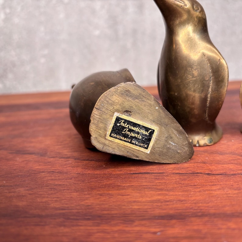 May include: A close-up of three small, vintage brass penguin figurines. One figurine is lying on its side, and the other two are upright. The label on the bottom of one reads "International Imports Handmade in Korea."