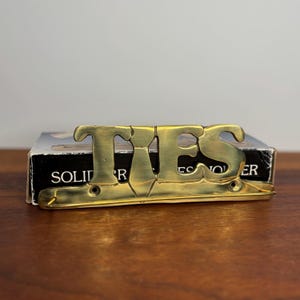 May include: A brass-coloured tie rack with the word "TIES" in large, stylised letters. The rack is mounted on a wooden surface, with a partially visible box behind it. The box has the word "SOLID" printed on it.