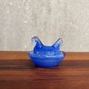 Könnte beinhalten: Eine kleine, durchscheinende blaue Glasfigur einer Henne, die in einem Nest sitzt. Die Henne hat eine strukturierte Oberfläche und sitzt in einer runden Schale mit gewelltem Rand, die das Aussehen eines geflochtenen Nestes aus blauem Glas nachahmt.