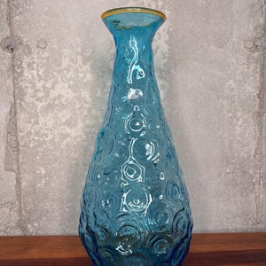 Puede incluir: Un jarrón alto de vidrio turquesa con un patrón texturizado similar a burbujas. El jarrón tiene una parte superior acampanada con un borde delgado de color dorado. El jarrón se exhibe sobre una superficie de madera contra una pared gris texturizada.