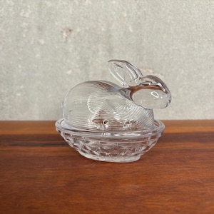 May include: A clear glass bunny figurine sits atop a basket-shaped dish. The bunny has detailed fur-like texture and long ears. The dish has a textured, basket weave pattern. The figurine is on a wooden surface.