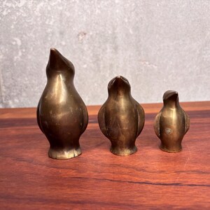 May include: Three antique brass bird figurines of varying sizes. The birds have a smooth, polished finish and are posed in an upright position. The figurines are displayed on a wooden surface against a neutral background.