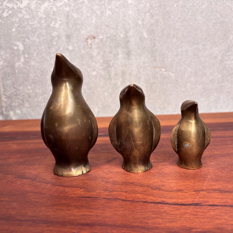May include: Three antique brass bird figurines of varying sizes. The birds have a smooth, polished finish and are posed in an upright position. The figurines are displayed on a wooden surface against a neutral background.