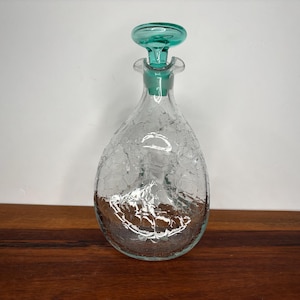 May include: A clear glass decanter with a crackle finish and a teal-colored stopper. The decanter has a teardrop shape and sits on a wooden surface. The stopper is round with a flared base.