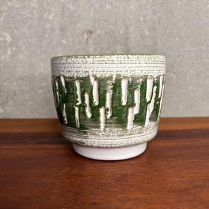 May include: A small ceramic bowl featuring a textured exterior with a white and dark green color scheme. The surface displays a horizontal brushed pattern layered over vertical, elongated white cutouts that reveal the base color underneath.